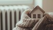 © Olga - Closeup of a small model house wrapped in a warm woolen scarf above a white radiator. Cold protection, home insulation, space heating system, energy crisis concept.