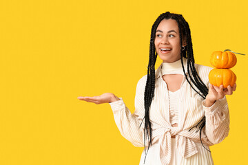 Poster - Beautiful young happy African-American woman with pumpkins pointing at something on yellow background