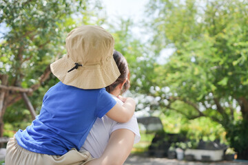 Asian mother is carrying her son on her back. The child is wearing a blue shirt and a hat.