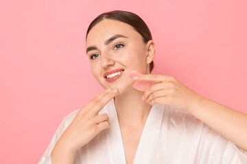 Wall Mural - Beautiful young woman doing facial massage with gua sha tool on pink background