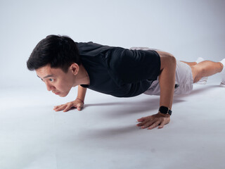 An Asian man performing a push-up in a studio setting. He is wearing a black t-shirt, gray shorts, and athletic shoes, determination during his fitness workout on a neutral light background.