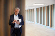 © Johnér - Senior businessman using mobile phone while standing against wall in office