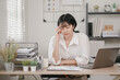 © phaitoon - A woman is sitting at a desk with a laptop and a stack of papers. She is wearing glasses and she is in a state of distress
