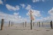 © Milou Dirks - young woman in long white dress and holding thin fabric in the wind standing on wooden poles near ocean on beach