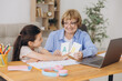 © anatoliycherkas - Happy grandma helping adorable preschool granddaughter school kid with homework task. Positive school pupil girl and granny reading learning book at laptop together, enjoying education at home