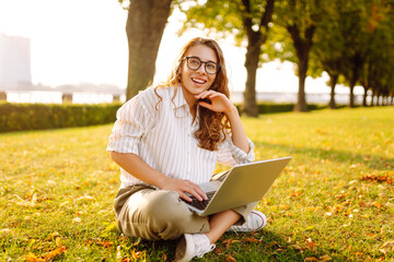 Wall Mural - Young woman working on a laptop outdoors in a sunny park during the afternoon, enjoying nature and the warm weather. Online education, Freelance work, technology concept.