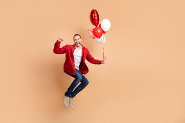 Full length portrait of nice young man jump balloons raise fist wear red shirt isolated on beige color background