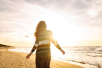 Wall Mural - Young woman enjoying a sunset at the beach while wearing a cozy striped sweater, smiling as waves gently lap at the shore. Travel, weekend, relax and lifestyle concept.