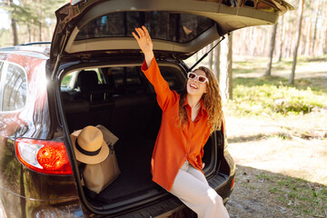 A traveler young woman smiles while sitting in the trunk of her car during a sunny day in a forested area with lush greenery. Road trips summer holidays.