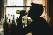© alfi - Silhouette of a man drinking from a bottle with empty and full glass bottles on a table, set in a softly lit somber scene