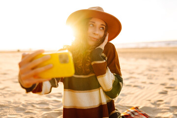 Wall Mural - Young woman taking a cheerful selfie on a sandy beach during sunset while wearing a hat and a striped sweater. Technology and lifestyle travel trip and journey concept