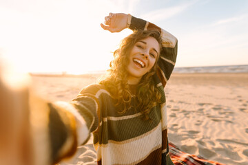 Wall Mural - Young woman taking a cheerful selfie on a sandy beach during sunset while wearing a hat and a striped sweater. Technology and lifestyle travel trip and journey concept