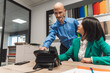 © VisualProduction - A business meeting scene featuring a man in a blue shirt and a woman in a green blazer. The man is pointing at a phone on the table while the woman looks at him attentively. The background shows color