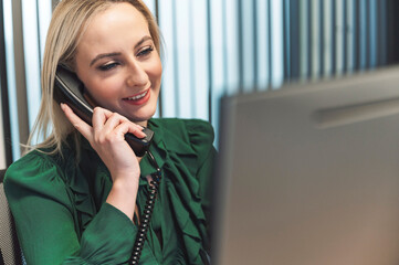 A woman in a green blouse is smiling while talking on the phone in an office setting. She is seated at a desk with a computer in front of her, surrounded by modern office decor.