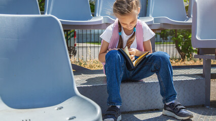 Elementary school girl or school pupil is studying, preparing to lesson, reading book or textbook outdoors at campus with backpack