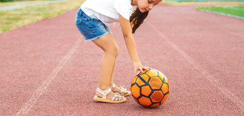 Child girl playing football at stadium. Little caucasian child ready to kick soccer ball at green field outdoors. Summer activities for children and future athlete concept.