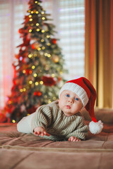 a baby boy in a santa claus hat on the background of a christmas tree. a baby in a knitted sweater.