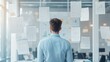 © sorrakrit - A businessman stands in a modern office, looking at a wall covered in papers and notes. He seems overwhelmed by the task ahead.
