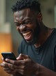© Alex - Black man joyfully laughing while engaged with smartphone in cozy indoor setting
