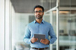© SejutaCahaya - A confident businessman in a blue shirt and glasses stands in a modern office holding a tablet.