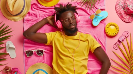 Wall Mural - Stunned dark skinned Afro American man in yellow t shirt and shorts lies on pink towel surrounded by sea accessories listens music headphones. Holiday, recreation, relax, Summer vibes