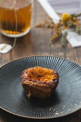 Wall Mural - Close-up of a traditional custard egg tart on a black plate with a cup of tea in the background