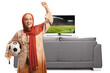 © Ljupco Smokovski - Woman in muslim ethnic clothes holding a football and cheering in front of tv and sofa