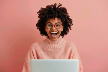 Cheerful young woman with an afro hairstyle and glasses, wearing a pink sweater, expressing excitement while using a laptop, set against a pink background.