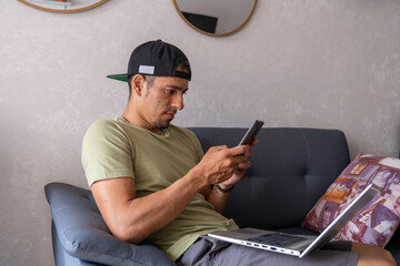 Un joven esta trabajando con su computador portatil y mirando el celular y sentado en el sofa de su casa.