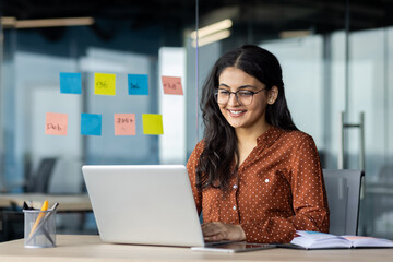 happy and smiling hispanic businesswoman typing on laptop, office worker with curly hair and glasses