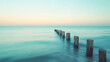 © Eddy Drmwn - A row of wooden posts extending into the calm, turquoise ocean at sunset.