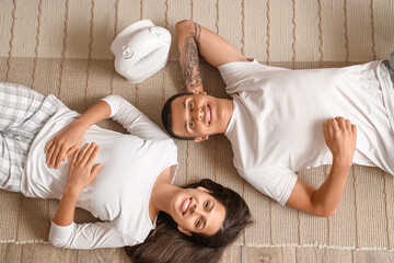 Wall Mural - Young couple with electric fan heater lying on floor, top view