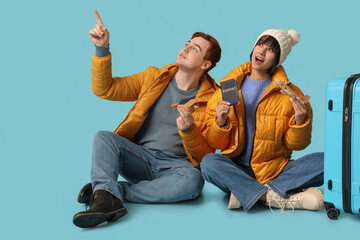 Wall Mural - Beautiful young shocked couple with suitcase, wooden airplanes and passport pointing at something on blue background. Winter vacation concept