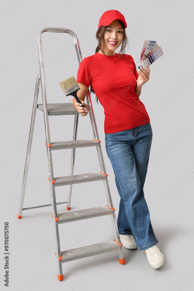 Young woman with paint brush, palette samples and ladder on grey background