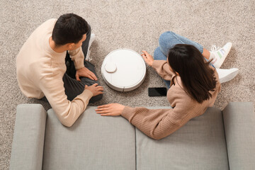 Poster - Young couple with modern robot vacuum cleaner and mobile phone sitting on floor in living room, top view