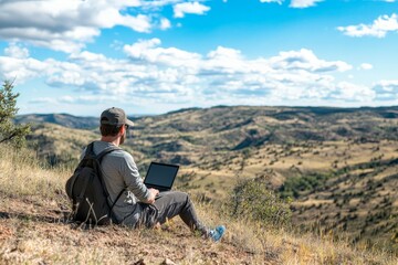 An adventurer immersed in his laptop work on a highland terrain with a stunning view of distant valleys, portraying remote work, freedom, and connection with nature.