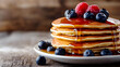 © Komkrit - Close-up of a stack of golden-brown pancakes drenched in maple syrup, topped with fresh blueberries and raspberries, with a rustic wooden background