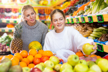 Portrait of friendly mother and teenage daughter making purchases together in greengrocery, choosing fresh fruits and vegetables