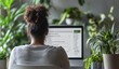© ShinneProject - A woman sits at her desk in front of her computer. She is surrounded by plants. Her back is to the camera, and she is looking at the computer screen.