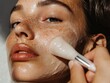 © Yotwarit - Close-up of a woman applying a facial cleanser with a brush.  Her face is covered in a white, foamy product.