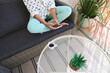 © Taras Grebinets - Woman relaxing on a patio sofa holding a smartphone with coffee and a plant on a wicker table for a peaceful outdoor setting