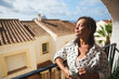 © Taras Grebinets - Woman enjoying a sunny morning on a balcony, feeling relaxed and peaceful while holding a cup of coffee, with charming Mediterranean architecture in the background.