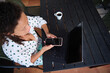 © Taras Grebinets - Woman using smartphone and laptop while working remotely on a patio with a cup of coffee, wearing a polka dot shirt.