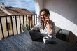 © Taras Grebinets - Smiling woman working on laptop during phone conversation at outdoor balcony with coffee and scenic views on a sunny day