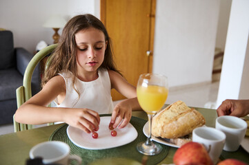 Young girl having breakfast at home with orange juice, bread, and grapes on a plate, enjoying a calm morning in a cozy dining room setting.