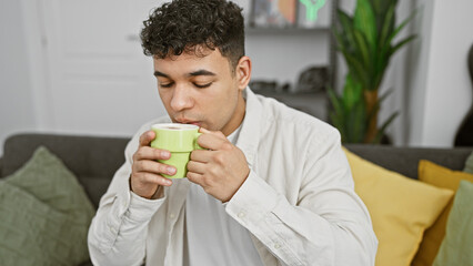 Wall Mural - A young man enjoys a warm drink at home, casually seated on a sofa in a modern living room with plants.