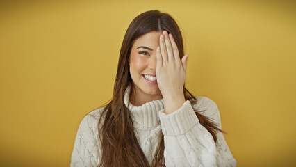 Wall Mural - A playful young woman in a white sweater covers one eye with her hand against a yellow background.