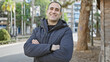 © Krakenimages.com - Smiling hispanic man with arms crossed wearing a jacket standing outdoors in a city park.
