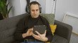 © Krakenimages.com - A relaxed man wearing headphones uses a tablet while sitting on a grey couch in a cozy living room.