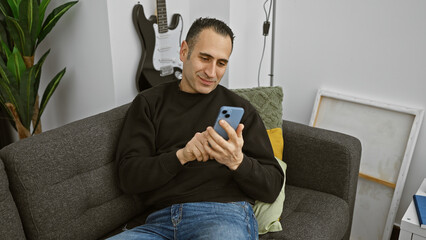 Handsome hispanic man sitting on a sofa in a cozy living room, engaging with his smartphone.